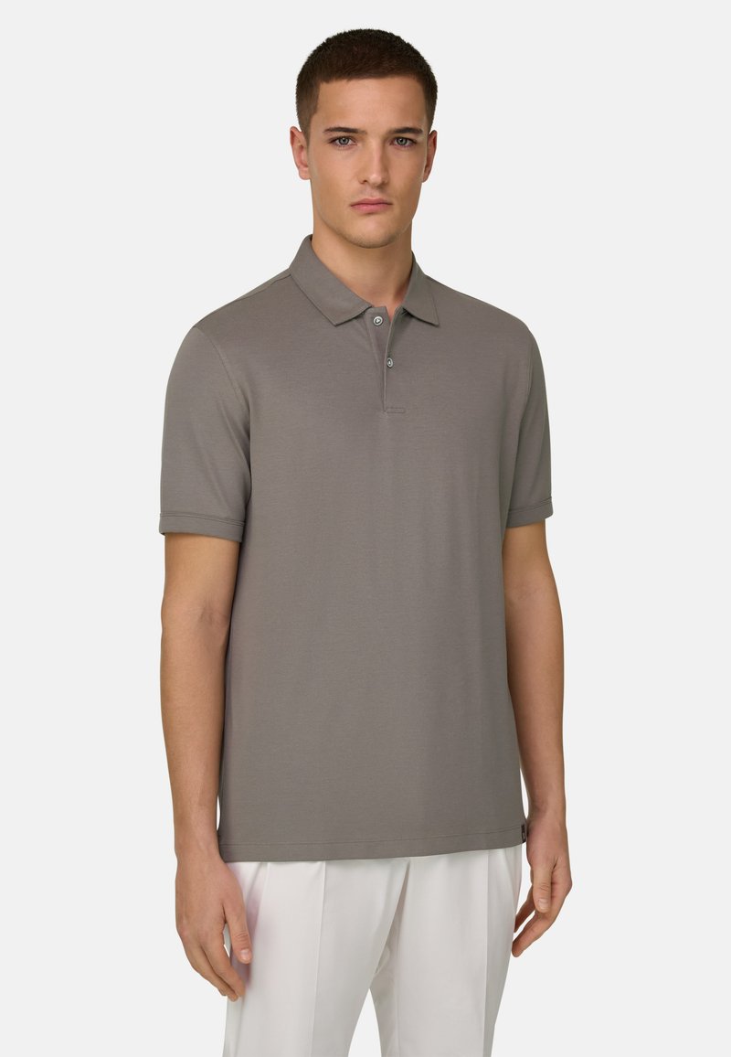 Homme portant un polo taupe à manches courtes avec deux boutons et un pantalon blanc, debout devant un fond clair uni.