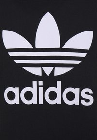 Juoda apranga su ryškiu baltu "Adidas" trefoil logotipu ir tekstu. Logotipas turi tris lapo formas ir tris horizontalius brūkšnius.
