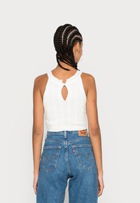 Top cropped de malha branca com um design de abertura nas costas, combinado com jeans de cintura alta azul com costura característica e um patch de logo em couro.