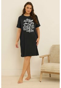 Kobieta stoi boso obok beżowego krzesła, ubrana w czarną sukienkę do kolan z białym napisem "Slow Sunday Coffee Club" i ilustracjami rogalików.