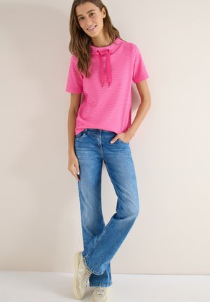 Jeune femme souriante, portant un sweat à capuche rose à manches courtes, un jean bleu et des baskets beige, debout devant un fond uni.