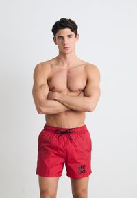 Shorts de bain rouges avec un cordon noir, arborant un logo "HUGO" sur la jambe gauche. Matériau léger et lisse adapté aux activités aquatiques.