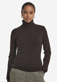 Kobieta w ciemno-brązowym swetrze z golfem i spodniach w wzór jodełki, z rękami w kieszeniach, nosząca złote kolczyki w kształcie kół, patrząca przed siebie.