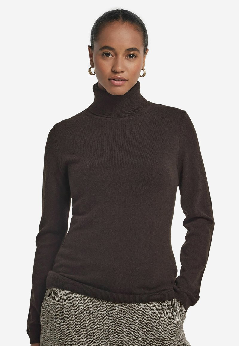 Kobieta w ciemno-brązowym swetrze z golfem i spodniach w wzór jodełki, z rękami w kieszeniach, nosząca złote kolczyki w kształcie kół, patrząca przed siebie.