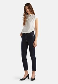 Bílá bezrukávová blůza s detailním skládáním u výstřihu, spárovaná s tmavě modrými úzkými džíny a černými lodičkami s špičatou špičkou.