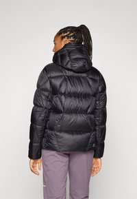 Cilvēks ar pinumiem matos valkā pūkainu melnu jaku, izgriezies prom, un apģērbts gaiši violetos biksēs, uz neitrāla fona.