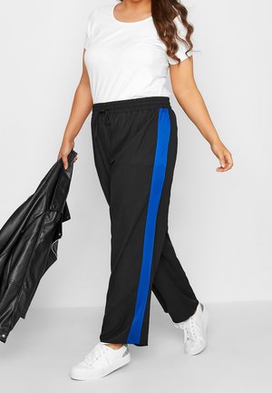 Frau trägt schwarze Hose mit blauem Seitenstreifen, weiße Sneakers und weißes T-Shirt, hält schwarze Lederjacke in der linken Hand.