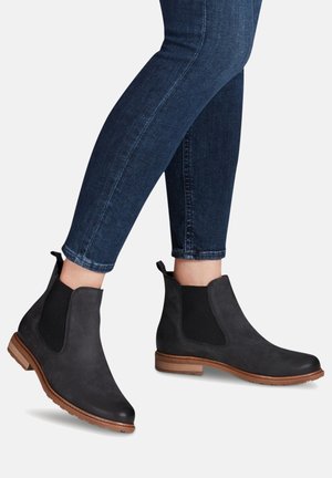 Schwarze Ankle Boots mit einem glatten Lederfinish, elastischen Seitenteilen und einem niedrigen braunen Holzabsatz, kombiniert mit enganliegenden dunklen Jeans.