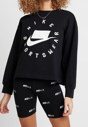 Osoba nosząca czarną bluzę Nike Sportswear z białym logo oraz czarne spodenki Nike Air z powtarzającym się białym napisem logo.