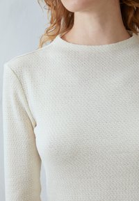 Vaalean beige neulepaita, jossa on textuurikuvio, pyöreä pääntie ja pitkät hihat. Paita esittelee yksinkertaista, vartalonmyötäistä muotoilua.