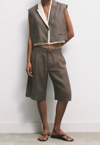 Chaqueta de traje marrón sin mangas y cortada, con pantalones cortos a la altura de la rodilla a juego, y chanclas negras en una persona de pie frente a una pared blanca.