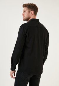 Maglietta nera a maniche lunghe con colletto, polsini con bottone e orlo dritto. Realizzata in un materiale liscio, presenta un design semplice.