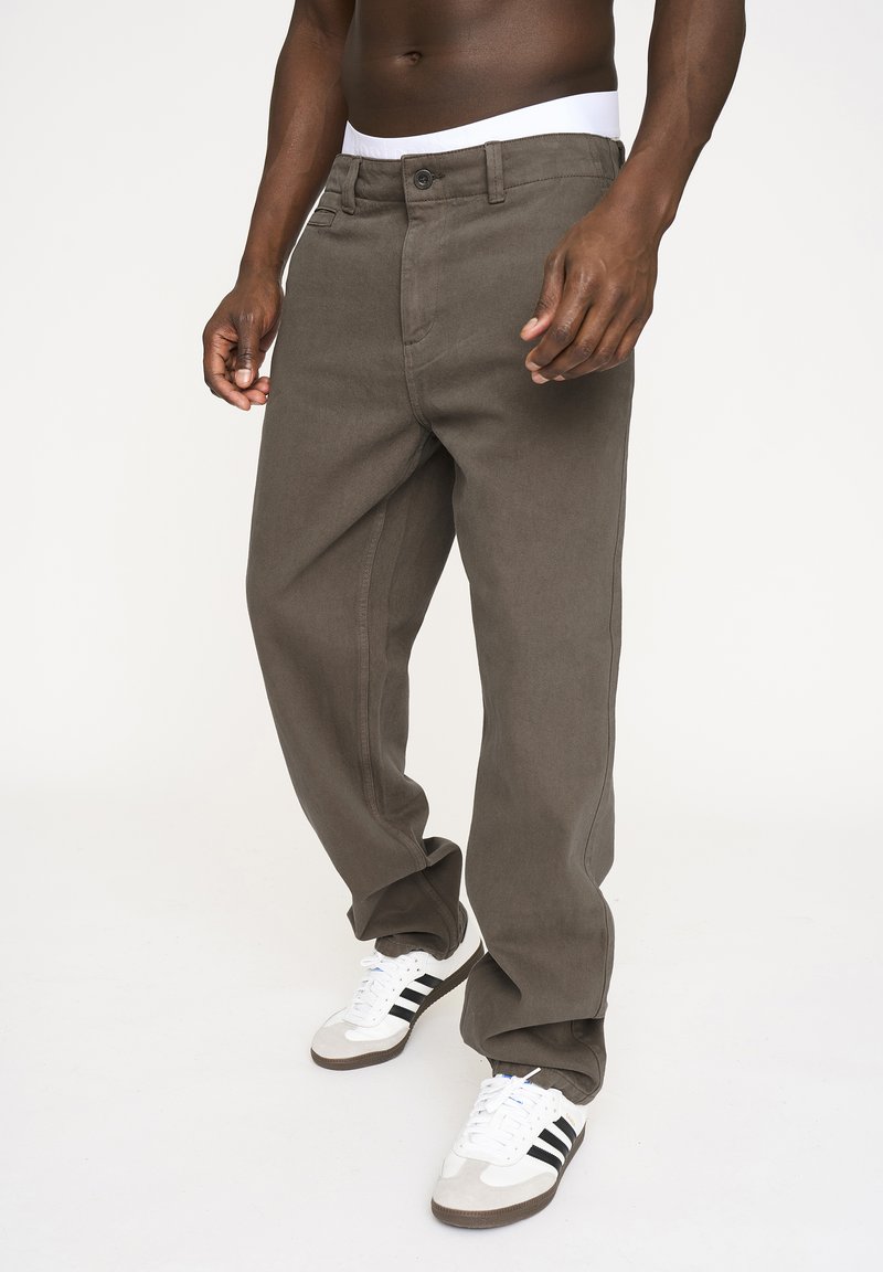 Bukser i mørke olivengrøn bomuldslerret, lige ben-design, med forlommer og knaplukning, båret med hvide og grå sneakers.