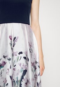 La robe présente un haut noir ajusté et une jupe fluide à motif floral dans des tons pastel doux, avec une texture lisse et un design plissé.