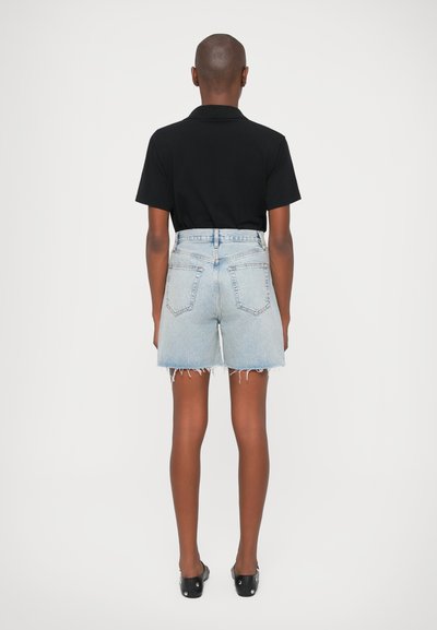 Fekete póló, világoskék farmer sorttal párosítva, amelynek szélén rojtosítás van és magas derékkal rendelkezik. A modell fekete lábbelije látható.