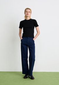 Sort tætsiddende T-shirt med korte ærmer sammen med marineblå vide bukser. Modellen har sorte sko med hvide striber på og står på et grønt tæppe.