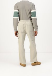 Osoba ubrana w jasnoszare spodnie cargo z elastycznym paskiem, beżowe sneakersy oraz longsleeve w zielono-białe paski na rękawach, stojąca.