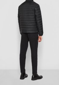 Veste en doudoune noire avec une texture lisse et matelassée, dotée d'un col montant et de poignets élastiques. Portée avec un jean noir slim et des bottines.