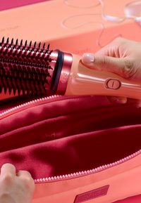 Main tenant une brosse à coiffure rose à côté d'une trousse de transport rose zippée ouverte sur une surface rose assortie.