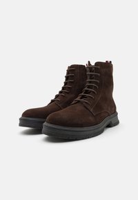 Un par de botas de tobillo de ante marrón oscuro con cordones, suelas de goma negra y cremalleras laterales sobre un fondo blanco.