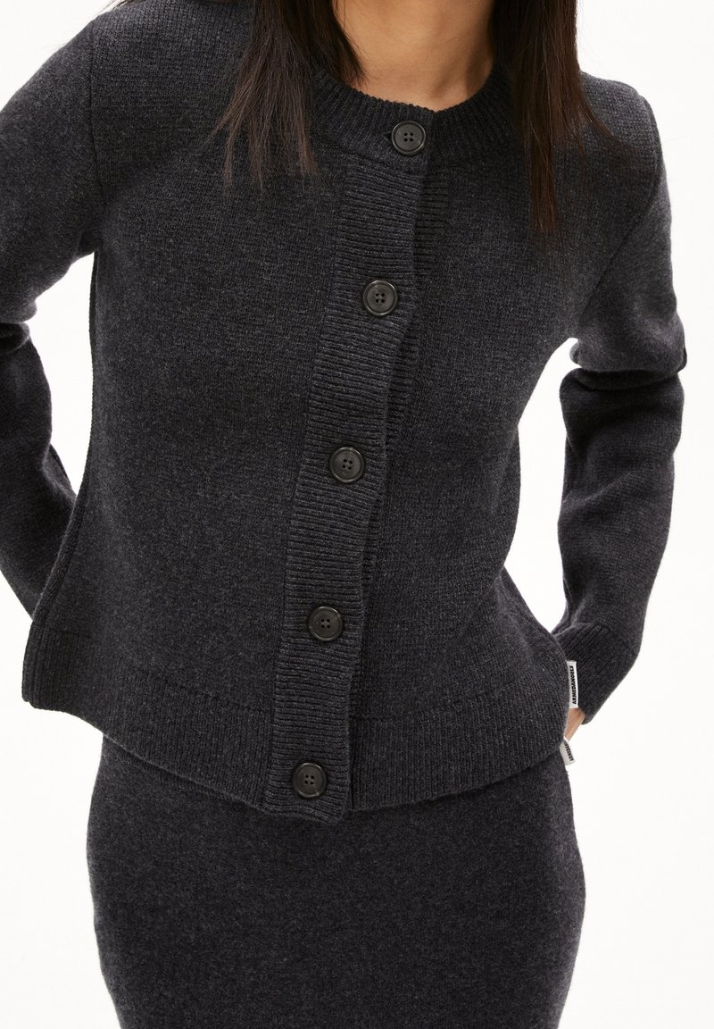 Femme portant un cardigan en maille gris foncé boutonné avec des manches longues et une jupe assortie, les mains placées derrière le dos.