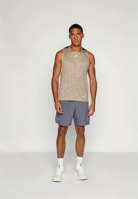 Top atletico color marrone chiaro con una texture sottile, abbinato a pantaloni corti grigi. Il modello è in piedi su uno sfondo neutro, indossando sneaker bianche.