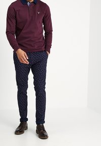 Man in een bordeauxrood poloshirt met lange mouwen, navy gekleurde slim-fit broek met patroon, en donkerbruin brogue schoenen, staand met één hand in zijn zak.