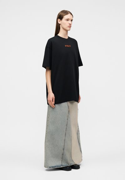 Γυναίκα που φοράει μια μαύρη μπλούζα μεγάλης διάστασης με το κείμενο "STIEGLITZ" και μια μακριά διπλή τόνου denim φούστα, στέκεται μπροστά σε μια απλή λευκή φόντο.