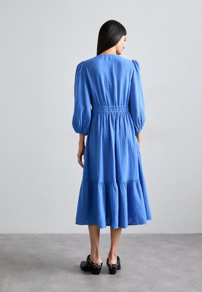 Blauwe jurk met lange mouwen, een smock taille en een tiered zoom. Gemaakt van lichtgewicht stof. Draagt een paar zwarte hoge klompen, zichtbaar vanaf de achterkant.