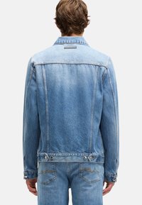 Spijkerjasje in lichtblauw. Voorzien van een klassieke kraag, knoopmanchetten, gestikte details en metalen accenten bij de taille. Achteraanzicht weergegeven.