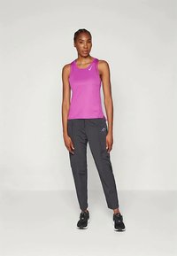 Γυναίκα όρθια που φοράει ένα αμάνικο ματζέντα αθλητικό τοπ, σκούρους γκρι tapered παντελόνες και μαύρα παπούτσια τρεξίματος σε απλή ανοιχτή φόντο.