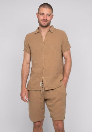 Homme aux cheveux courts et à la barbe, portant une chemise boutonnée à manches courtes marron clair et un short assorti, debout avec une main dans la poche.