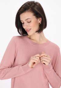 Világos rózsaszín, hosszú ujjú pulóver, puha textúrával és bordás nyakkivágással, egy személy viseli, aki összefonja a kezét.