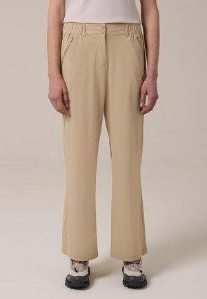 Personne portant un pantalon beige à jambes droites avec poches zippées et des chaussures de randonnée beiges, debout devant un fond uni.