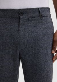 Grau gemusterte Hose aus strukturiertem Stoff, mit einem Knopfverschluss und einem kleinen schwarzen Akzent in der Nähe des Taillenbunds.