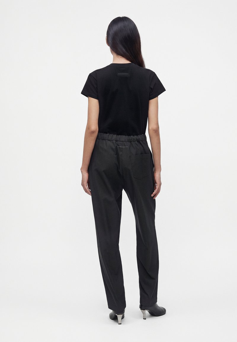 Femme aux longs cheveux noirs portant un t-shirt noir à manches courtes, un pantalon noir ample avec une taille élastique et des talons noirs, vue de dos.