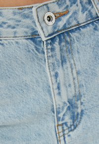 Vaqueros de mezclilla azul claro con una cintura alta, cierre de botón metálico y costuras en contraste. Tejido texturizado con sutiles detalles de desvanecimiento.