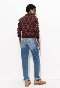 Pull en argyle marron et rouge avec des poignets côtelés, associé à un jean en denim bleu clair et des chaussures beiges avec des lacets marron. Fond minimaliste.