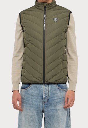 Homme portant un gilet matelassé sans manches olive avec des poches zippées par-dessus une chemise beige à manches longues et un jean bleu clair.