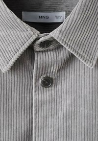 Camisa de pana gris con cuello puntiagudo, canalé vertical y botones negros en la tapeta. Etiqueta visible en el interior del cuello.