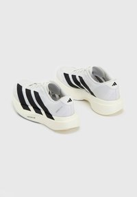 Paire de chaussures de course blanches avec des rayures noires et des semelles épaisses blanc cassé sur un fond clair, montrant les vues arrière et latérale.