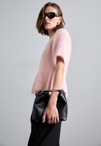 Růžový, chlupatý svetr s krátkým rukávem, doplněný černou skládací kabelkou se stříbrnou rukojetí. Model má na sobě tmavé kalhoty a sluneční brýle.