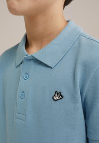 Gaiši zils polo krekls ar teksturētu audumu, trīs pogām un melnbaltu izšūtu putna logo kreisajā krūtī.