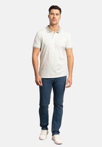 Polo de color gris claro con un patrón texturizado y cuello verde, combinado con jeans azul oscuro y zapatillas deportivas blancas.