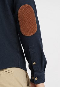 Marinblå långärmad skjorta med manchesterlappar vid armbågarna, knäppningsmanschetter och en strukturerad yta. Krage med en svag sväng vid nederkanten.