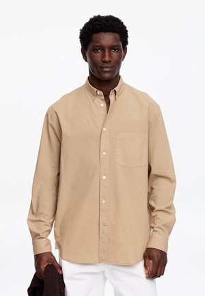 Man draagt een beige overhemd met borstzak en witte broek, houdt een donkerkleurig kledingstuk in één hand vast.