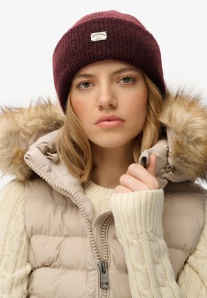 Bordeaux gebreide beanie met een label, beige puffer vest met nepbont, en een crèmekleurige kabelgebreide sweater. Close-up van de details van de kleding.