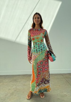 Γυναίκα που φοράει μακρύ, πολύχρωμο φόρεμα πατςγουορκ με σχέδια ήλιου και ματιού, κρατώντας τσάντα που ταιριάζει, στέκεται σε τσιμεντένιο δάπεδο δίπλα σε λευκό τοίχο.