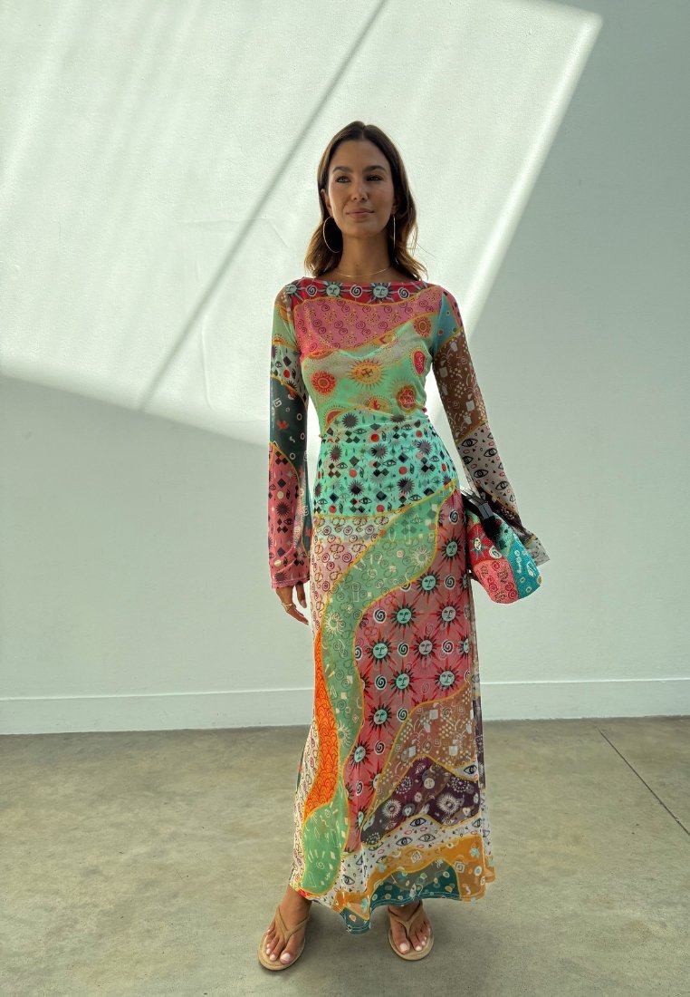 Γυναίκα που φοράει μακρύ, πολύχρωμο φόρεμα πατςγουορκ με σχέδια ήλιου και ματιού, κρατώντας τσάντα που ταιριάζει, στέκεται σε τσιμεντένιο δάπεδο δίπλα σε λευκό τοίχο.