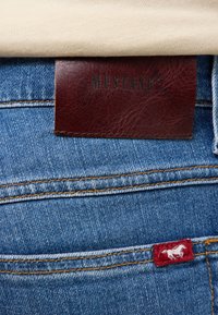 Niebieskie dżinsy z brązową skórzaną metką oznaczoną "MUSTANG" oraz czerwoną etykietą z białym koniem haftowaną na tylnej kieszeni.